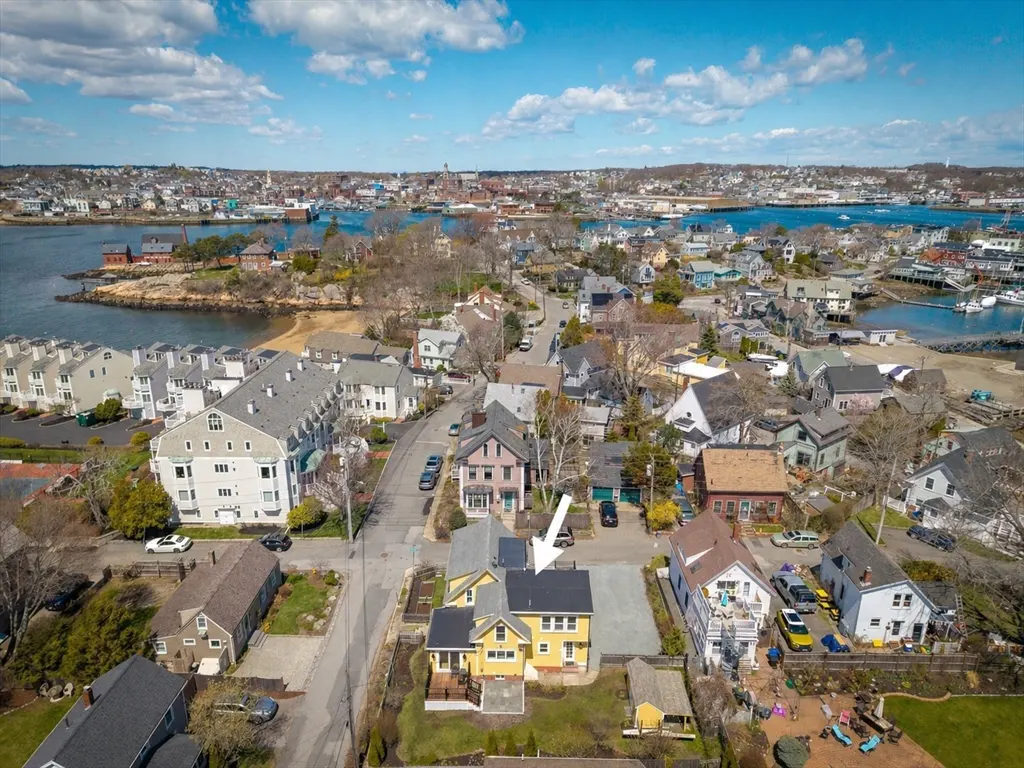 Aerial view of a yellow house with a black roof, surrounded by other houses and a body of water in the background.