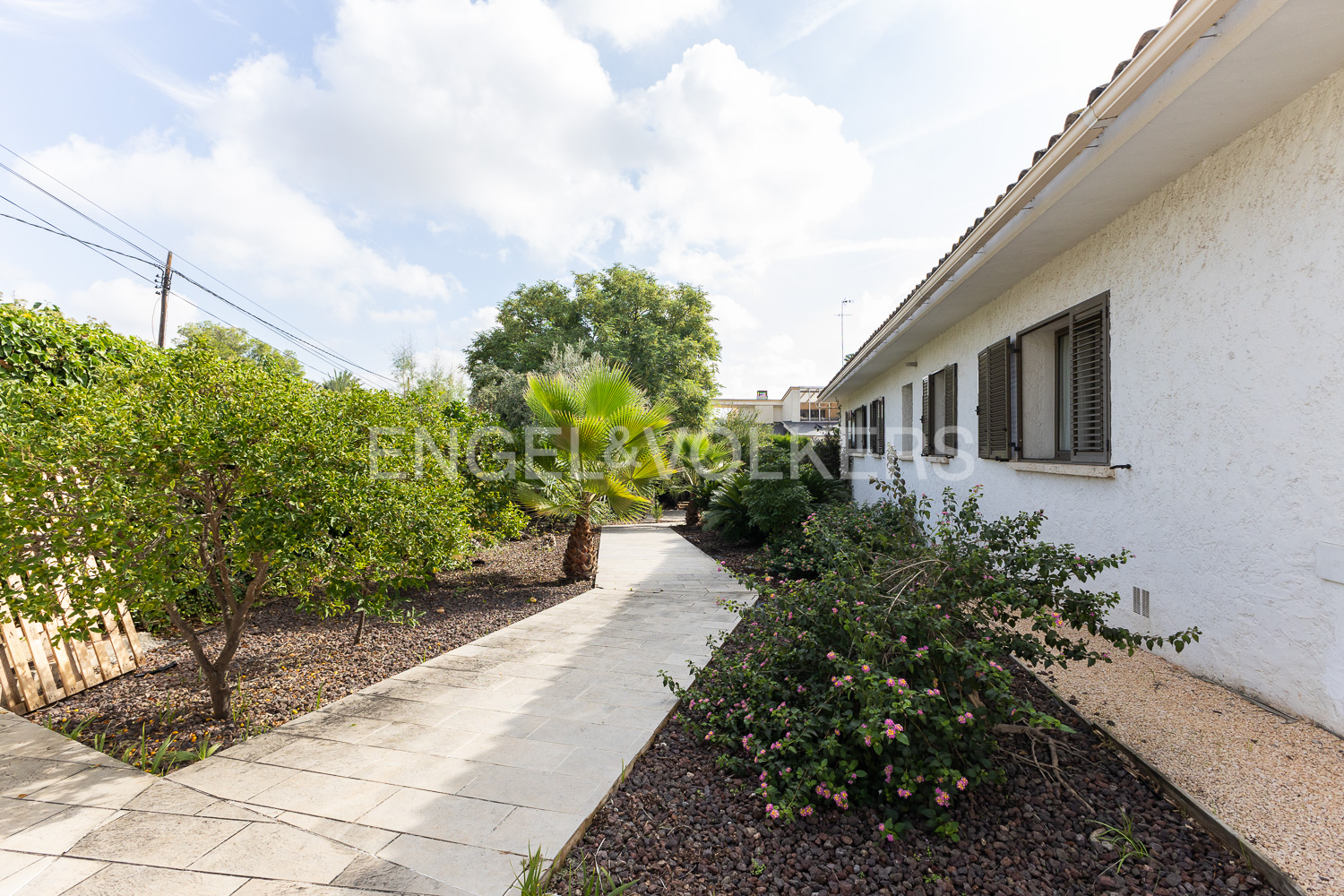 Exterior view of a white house with brown shutters, a stone walkway, and lush green landscaping under a partly cloudy sky.