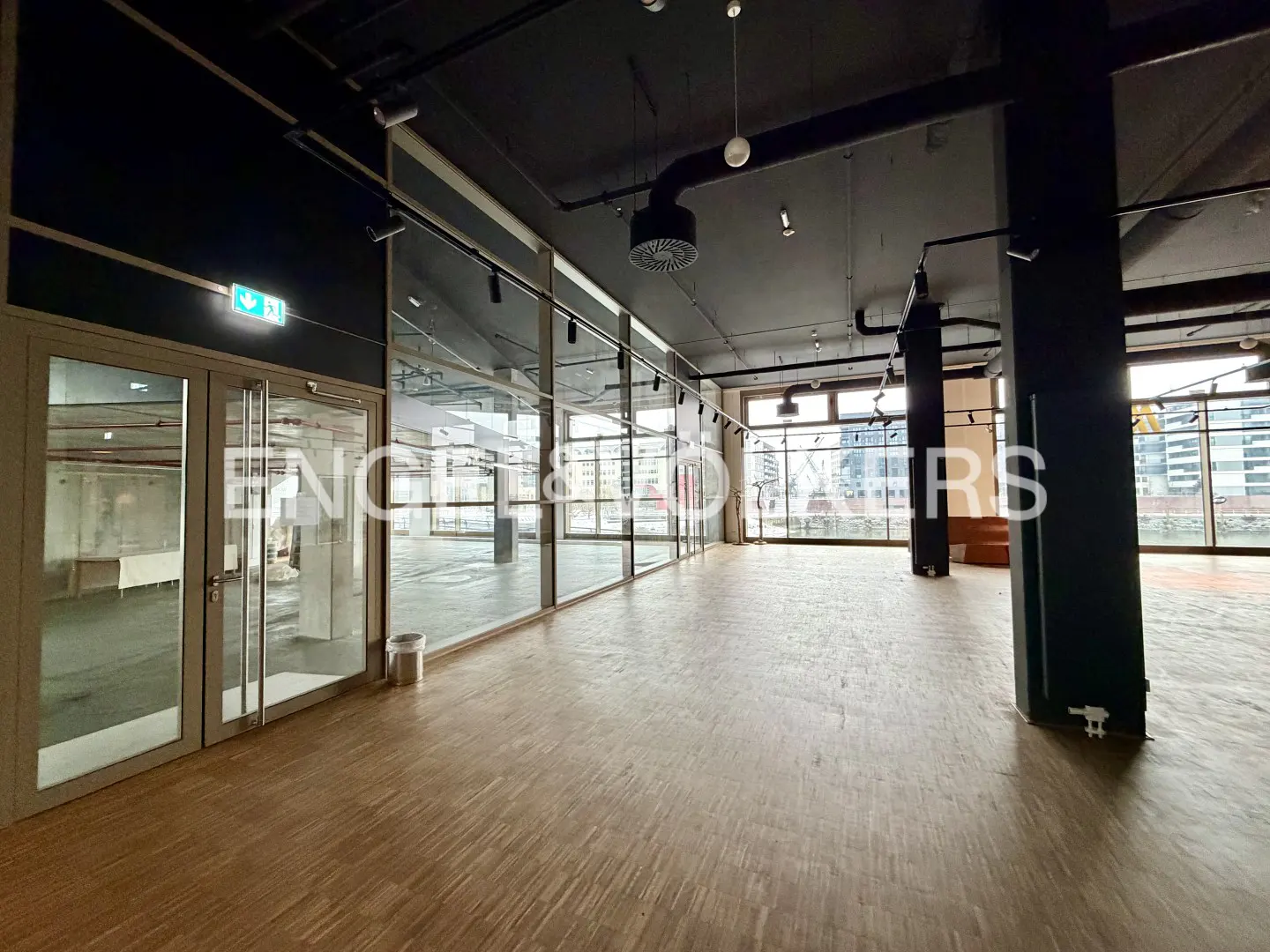 Empty commercial space with wood floors, black ceiling, and large windows. An exit sign is above the glass doors.