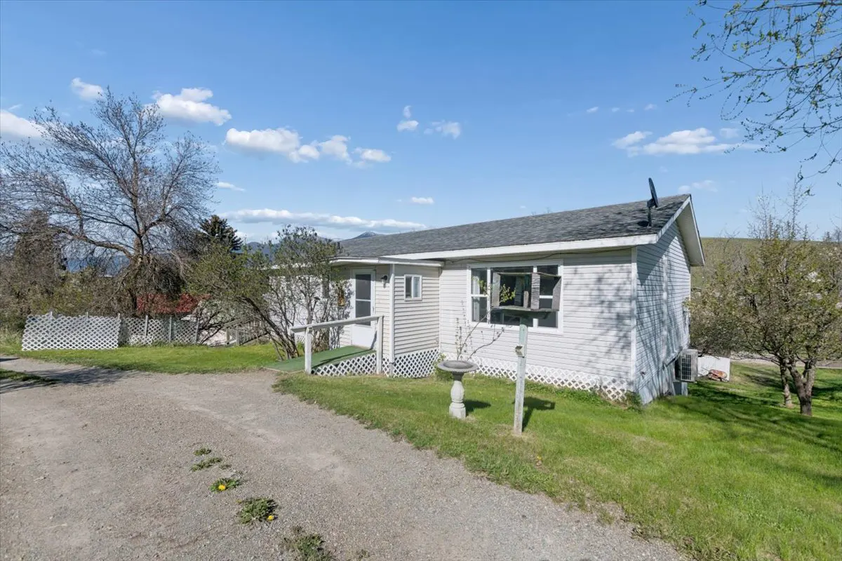A single-story, light gray house with a dark gray roof, green lawn, and gravel driveway on a sunny day.