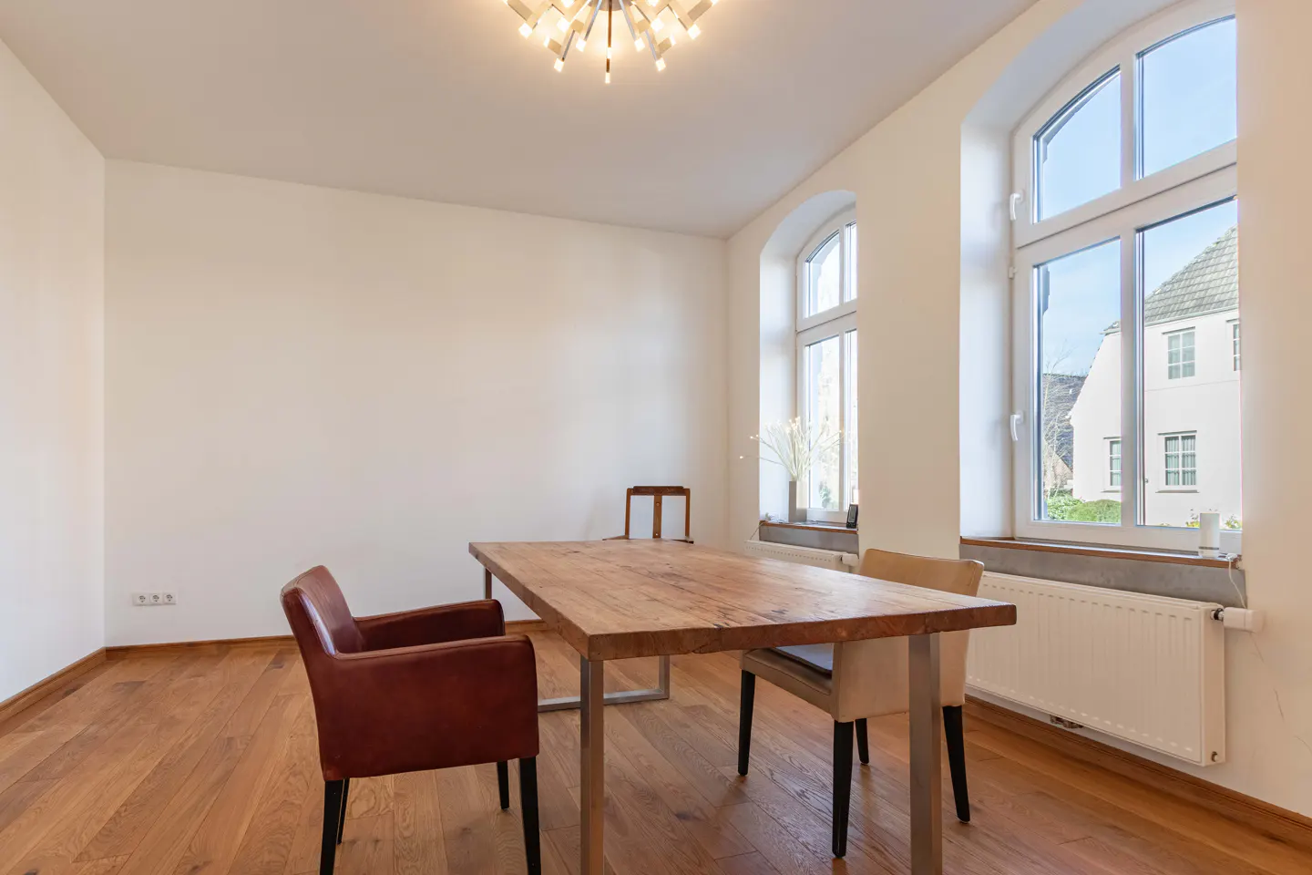 Bright dining room with wood floors, white walls, and a wooden table with metal legs. Two chairs are visible, one brown leather. Arched windows let in natural light.