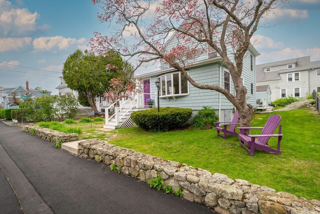A light blue house with a purple door and two purple Adirondack chairs on the lawn. A stone wall lines the street.