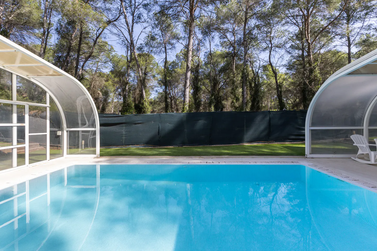 Outdoor pool with clear blue water, enclosed by white arched covers. Trees and a green fence are in the background.