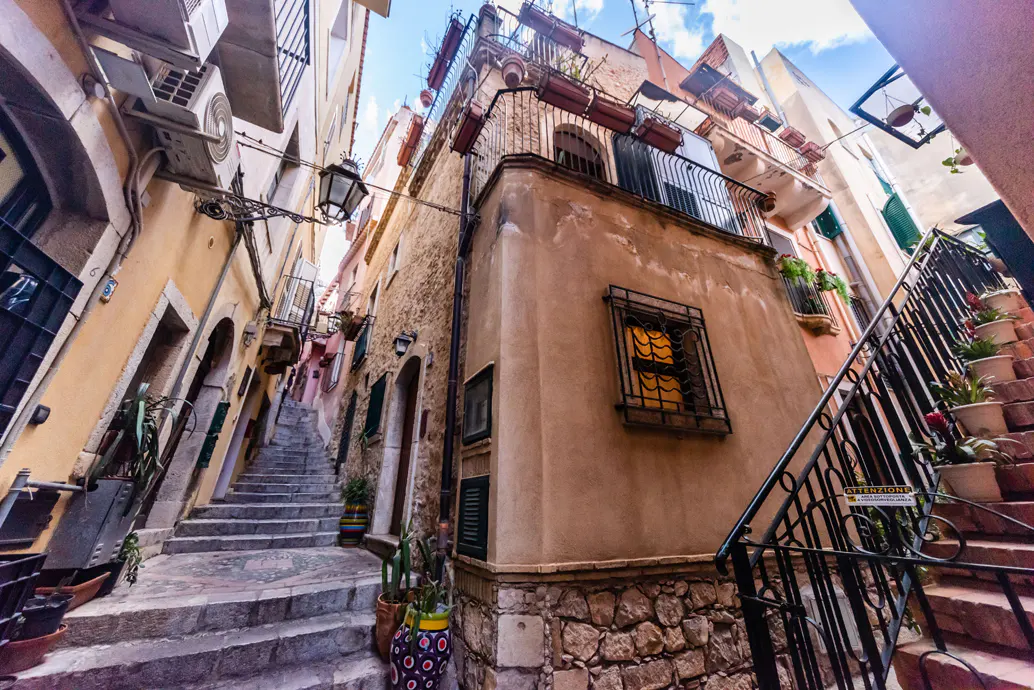 A narrow street view with stone steps, old buildings, and wrought iron balconies in Taormina, Italy.