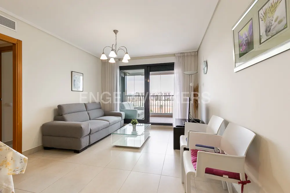Bright living room with a grey sofa, glass coffee table, two white chairs, and a balcony door with sheer curtains.