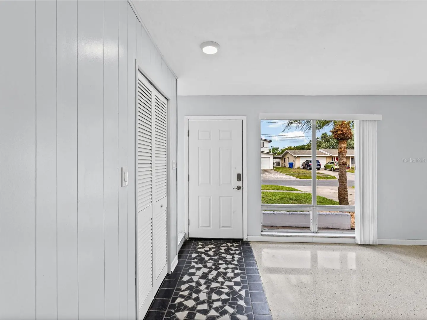 Entryway with white door, closet, and black and white tiled floor. A large window shows a palm tree and a suburban street.