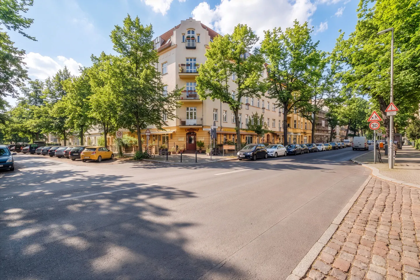 Street view of a light yellow corner building with a restaurant, cars parked along the street, and green trees.