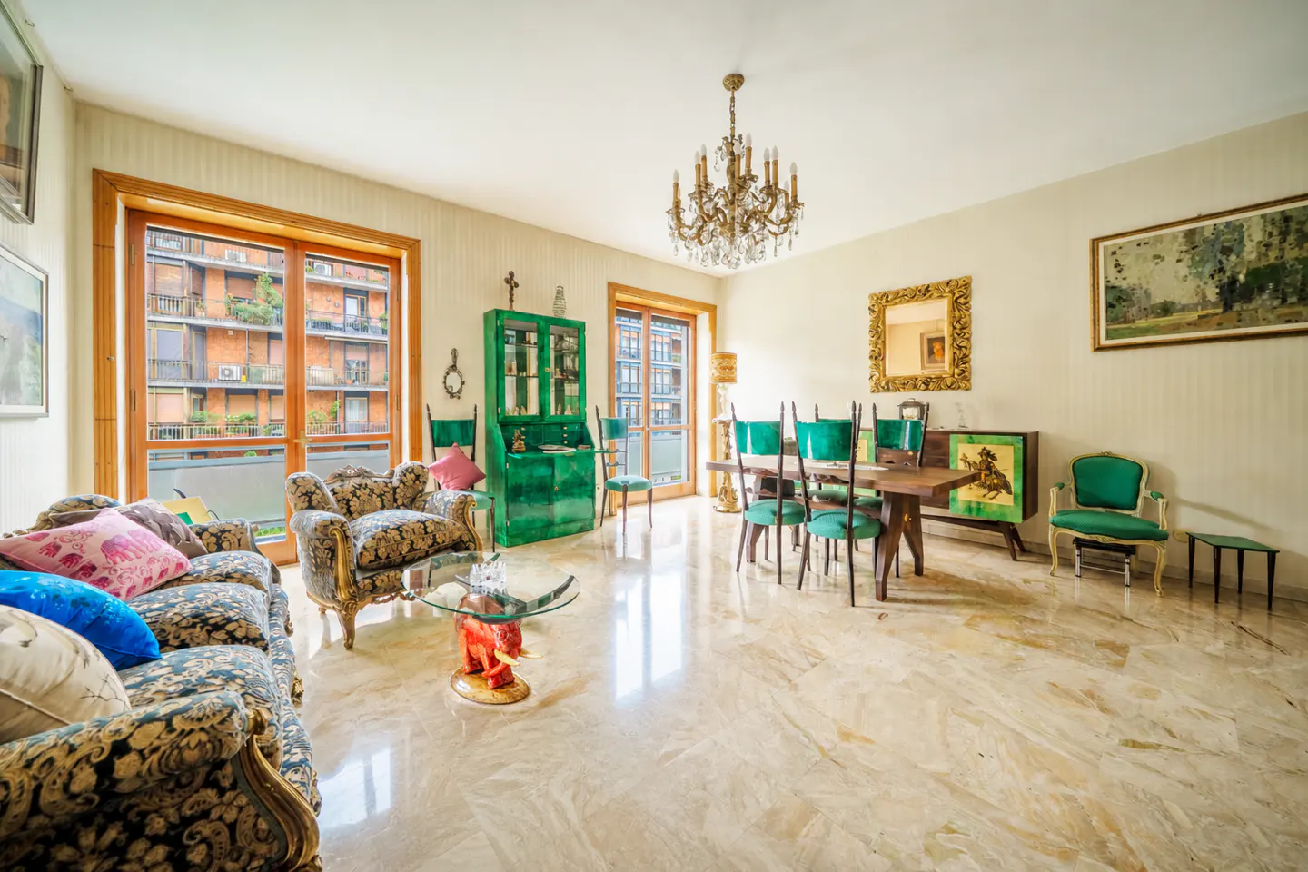 Bright living room with marble floors, floral sofa, glass table, and green dining set under a chandelier. Balcony view of a brick building.