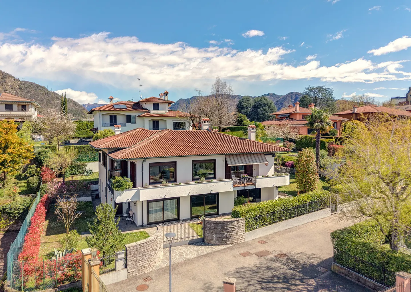 Two-story white house with a red tile roof, surrounded by lush greenery and a stone wall, under a blue sky with clouds.