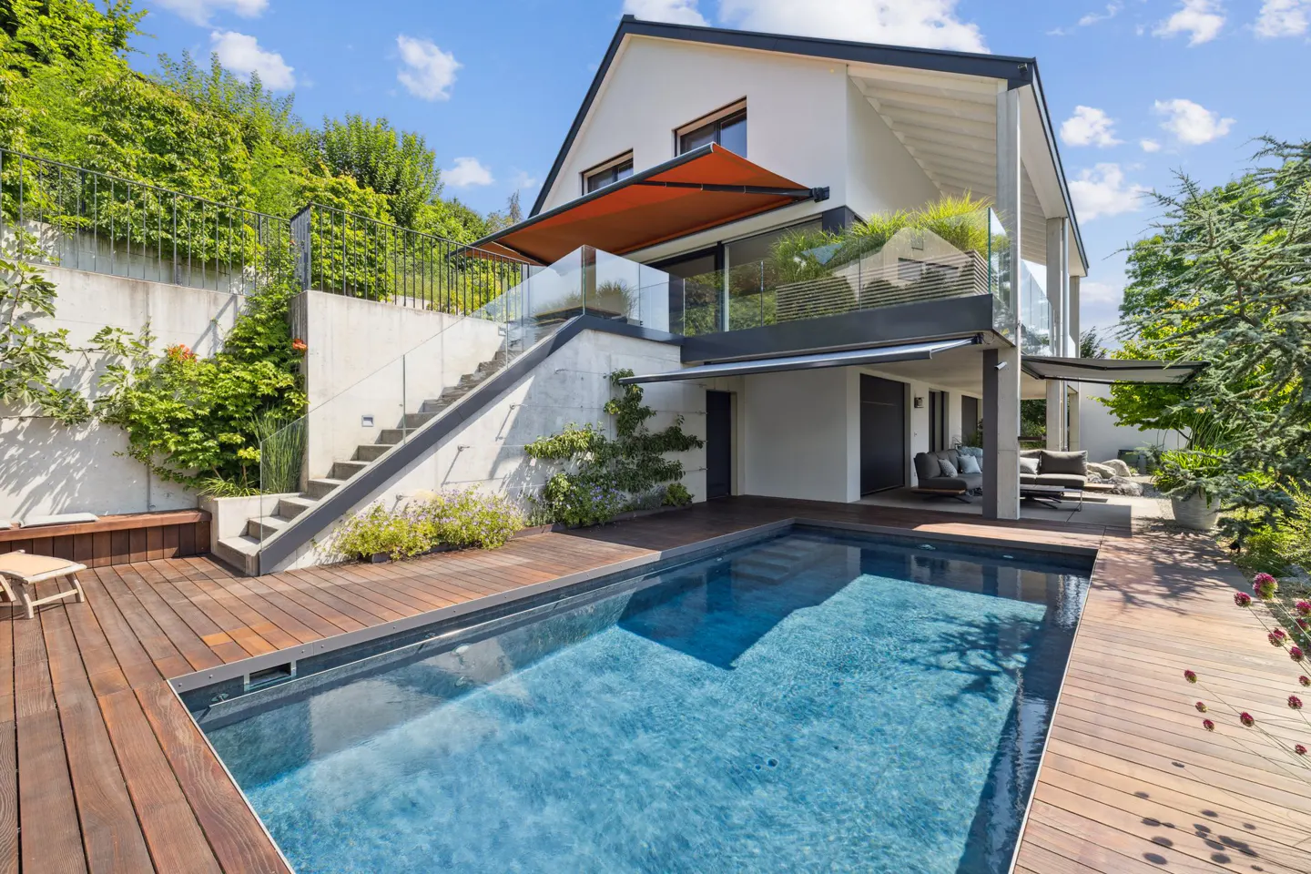 Modern white house with a blue swimming pool and wooden deck. Stairs lead to a glass-railed patio with an orange awning. Lush greenery surrounds the property.