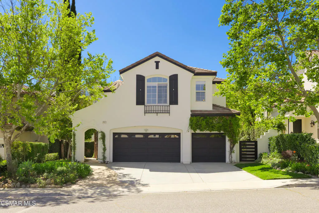 Two-story house with a white stucco exterior, dark brown roof, and a two-car garage. Green trees frame the house against a blue sky.