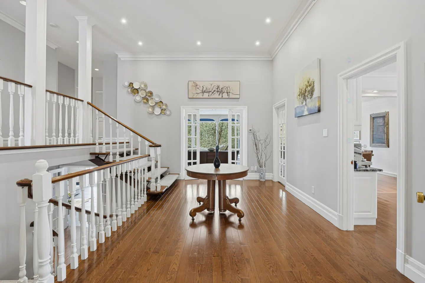 Bright foyer with hardwood floors, white staircase, and round table with vase. Art adorns the gray walls.