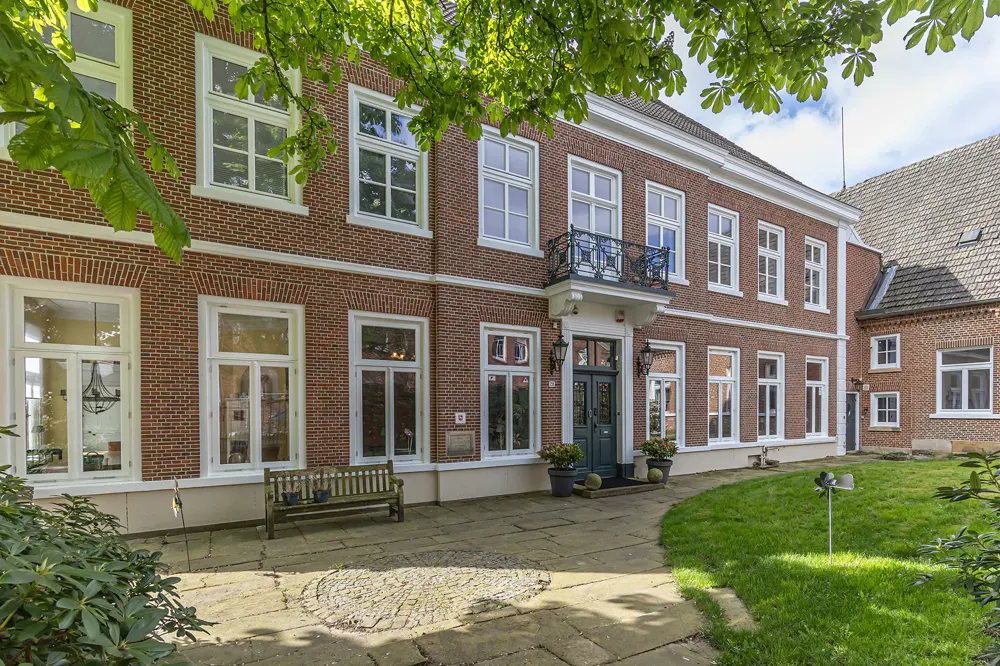 Exterior view of a two-story red brick building with white-framed windows and a small balcony above the entrance. A stone walkway leads to the front door.