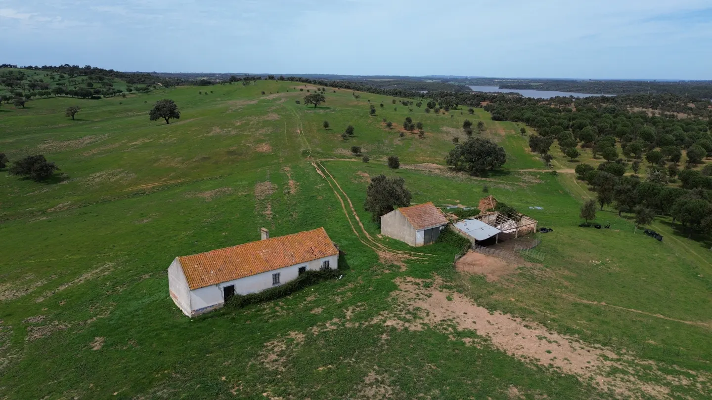 Aerial view of a rural property with a white house, orange roof, and outbuildings on a green hillside.