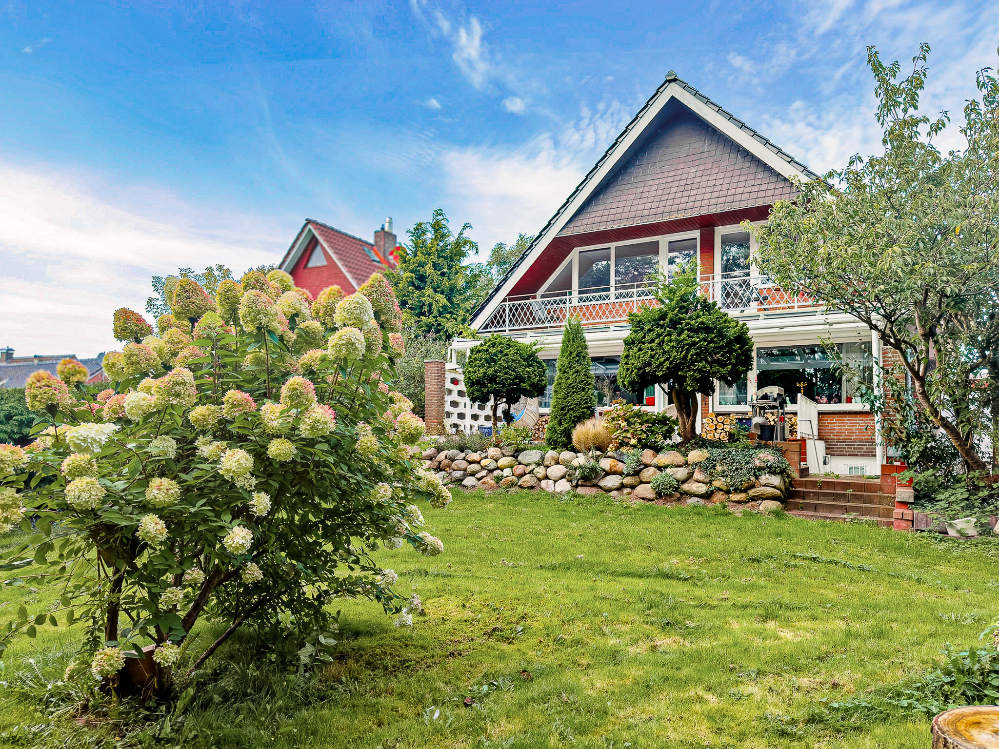 A red-roofed house with a balcony sits behind a lush green lawn, a large hydrangea bush, and a stone garden wall. [AI-generated text]