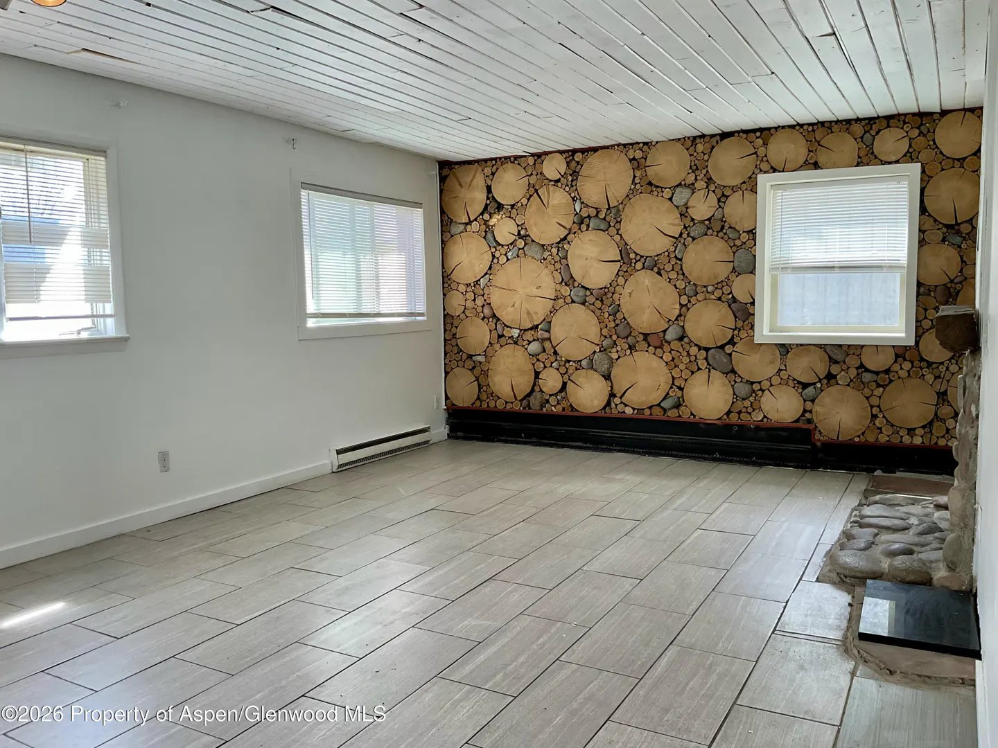 A bright room with white walls, wood-look tile floor, and a log-covered wall with a stone fireplace.