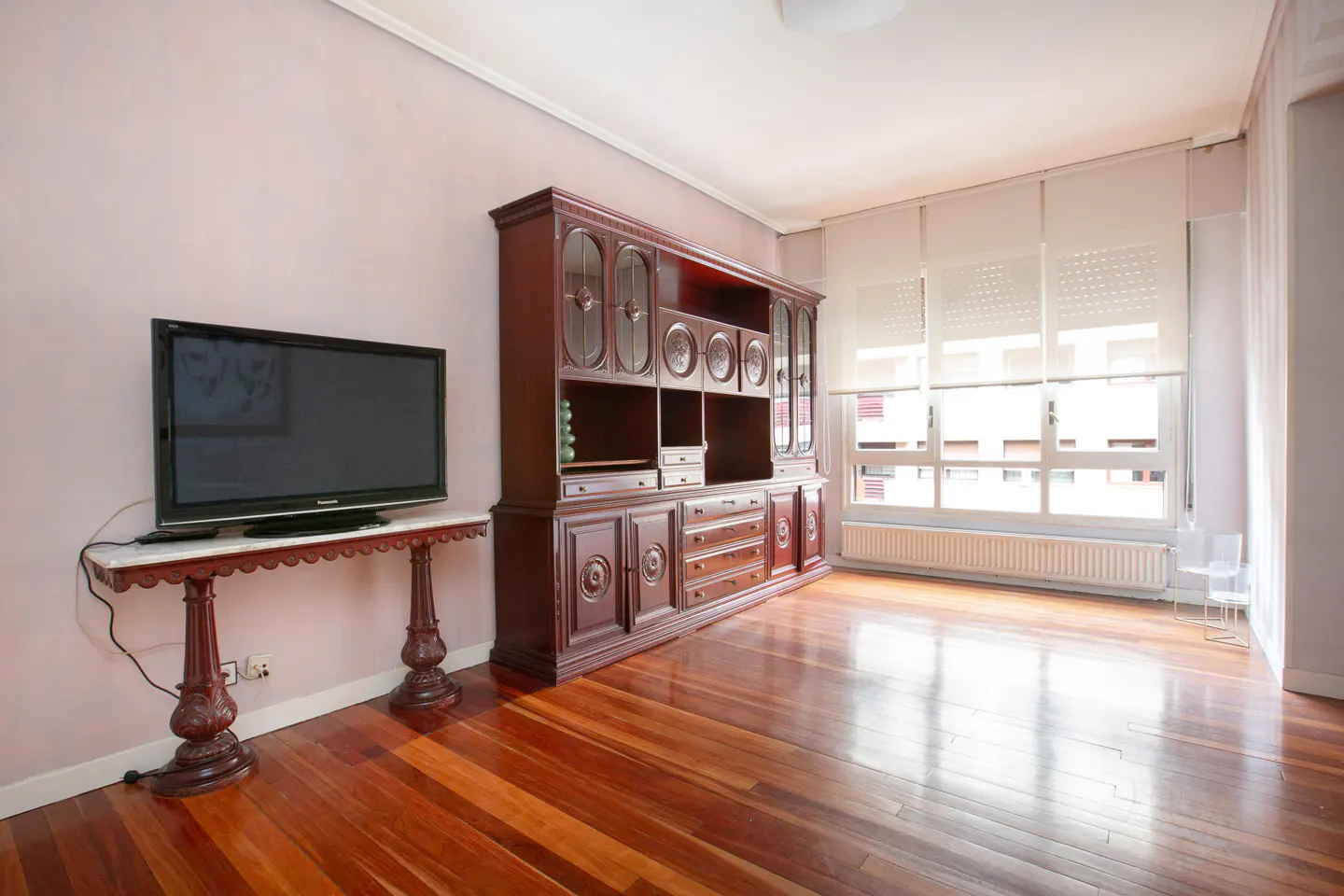 Living room with hardwood floors, a large dark wood cabinet, and a TV on a marble-topped table. Windows with blinds let in natural light.