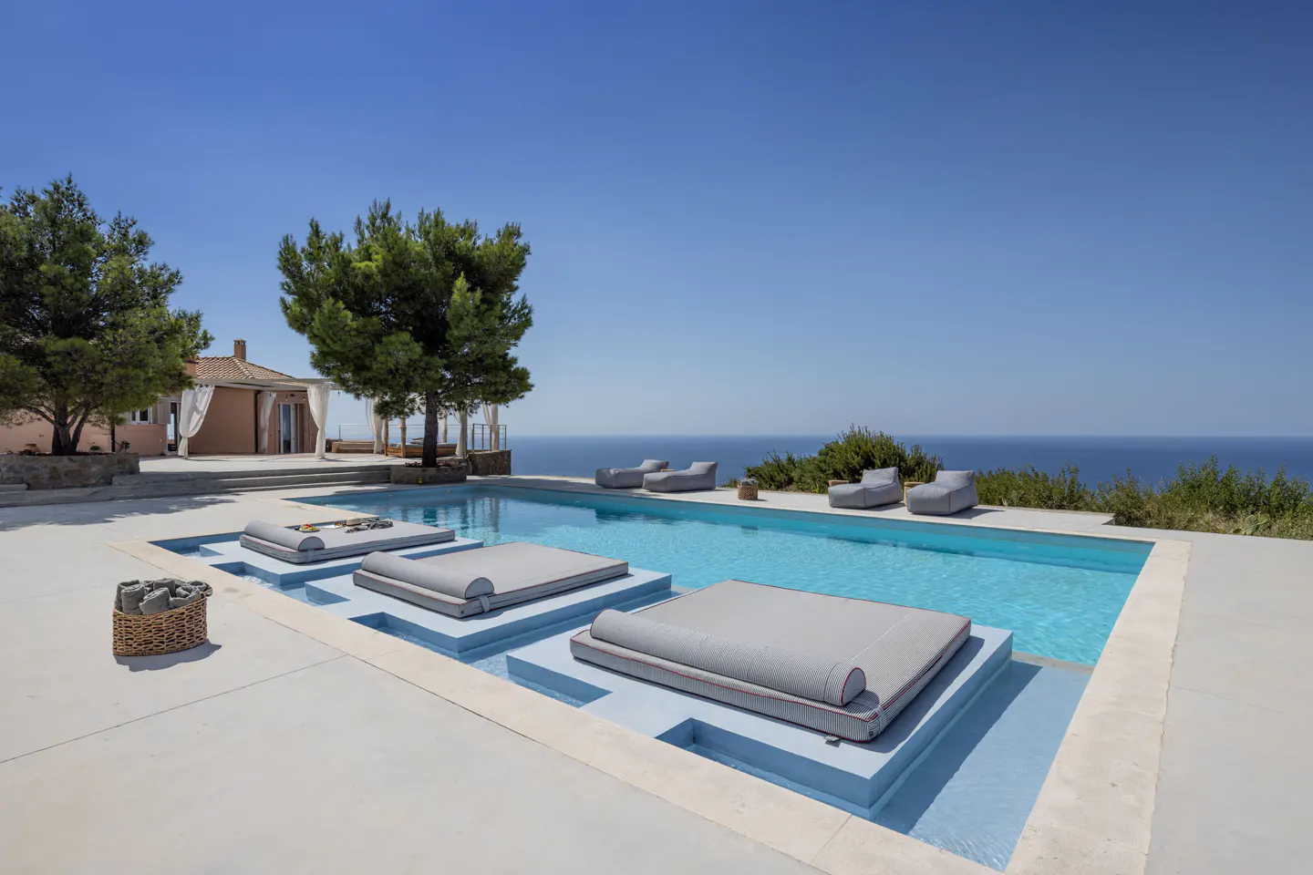 A luxury villa with a pool featuring built-in lounge beds and ocean views under a clear blue sky.