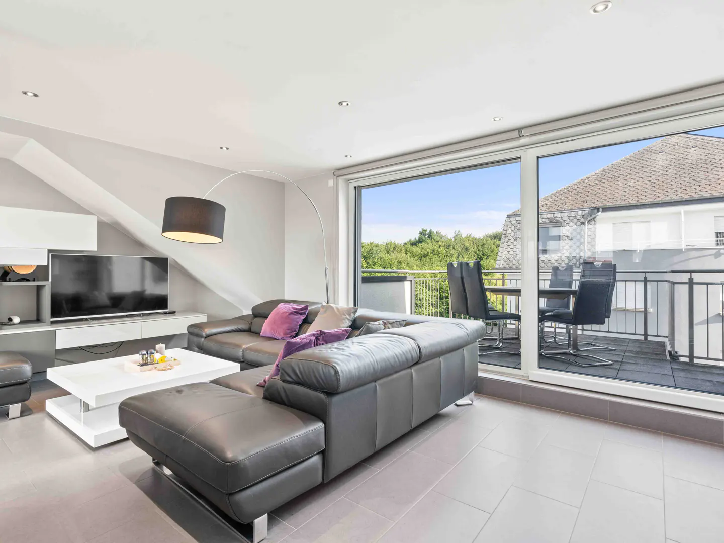 Living room with gray leather sectional, white coffee table, TV, and balcony with outdoor dining set.