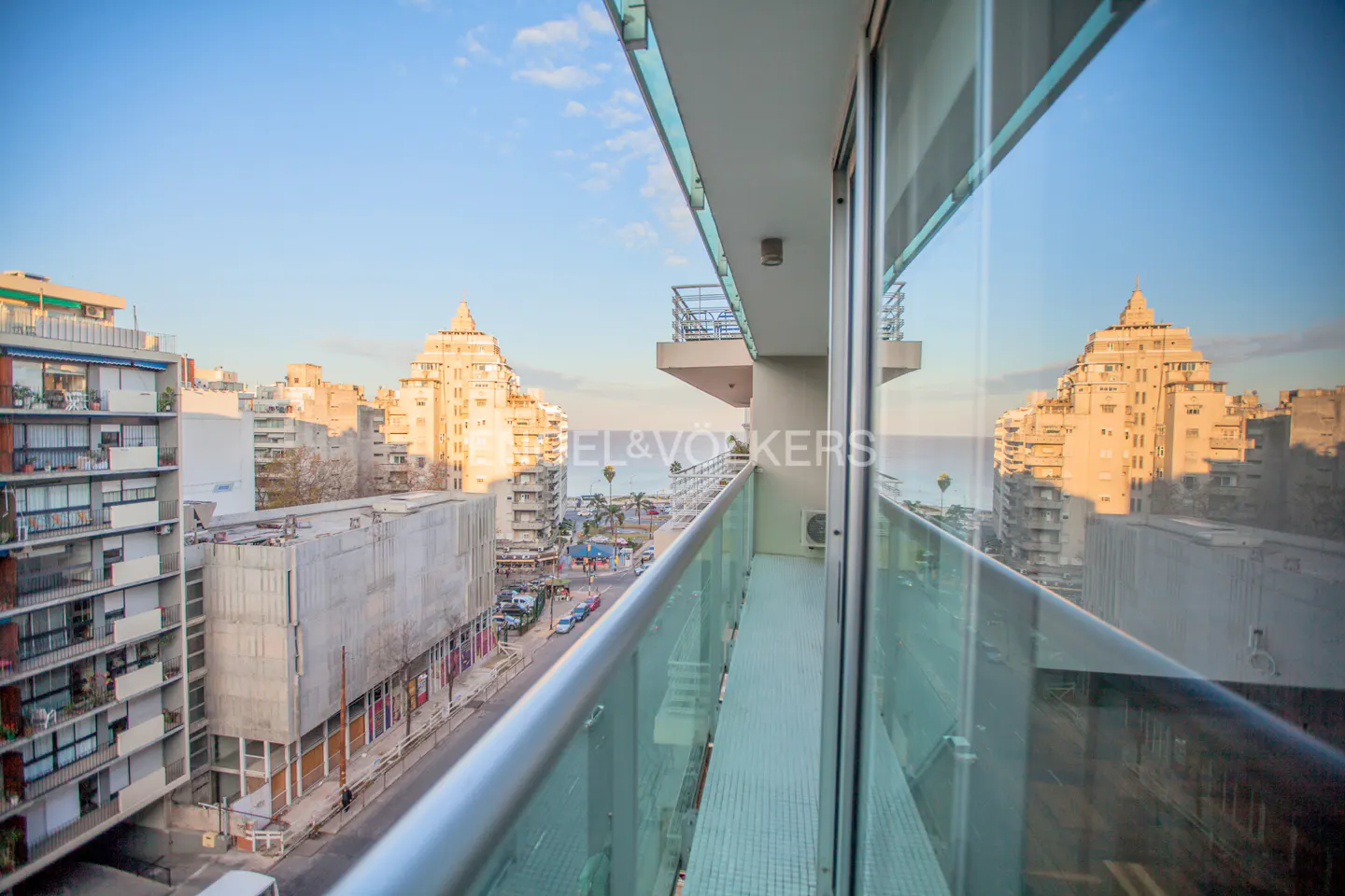 View from a balcony with glass railings overlooking a city street and the ocean. Buildings are reflected in the glass.
