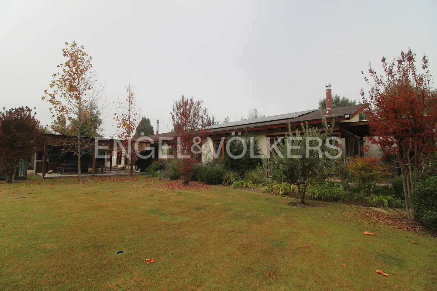 A single-story house with solar panels, a chimney, and a large green lawn with trees. The sky is overcast.