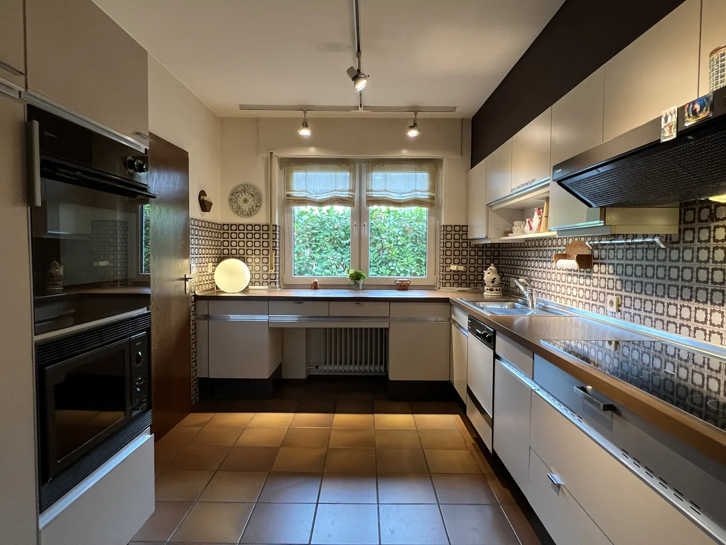 A kitchen with white cabinets, brown countertops, and brown and white patterned backsplash. A window looks out onto green foliage.
