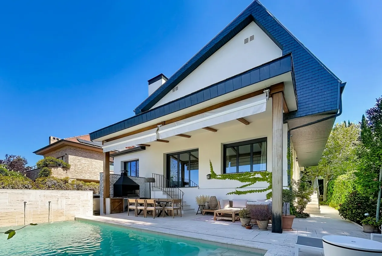 Backyard view of a modern white house with a blue roof, pool, patio furniture, and grill on a sunny day.