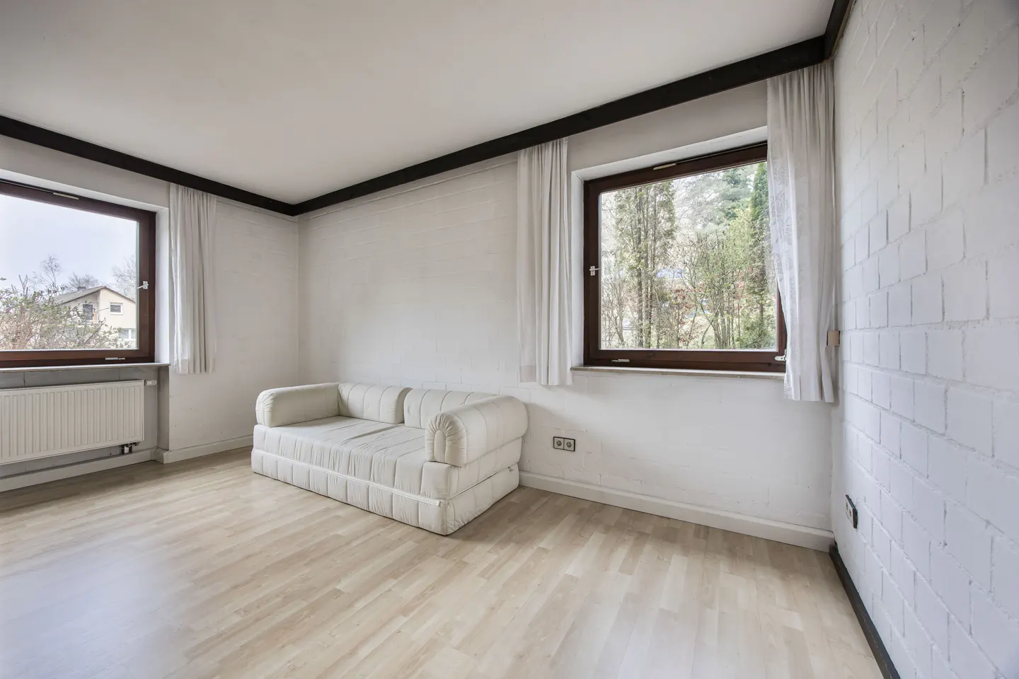 Bright, empty room with light wood floors, white walls, and a white sofa. Two windows with white curtains let in natural light.