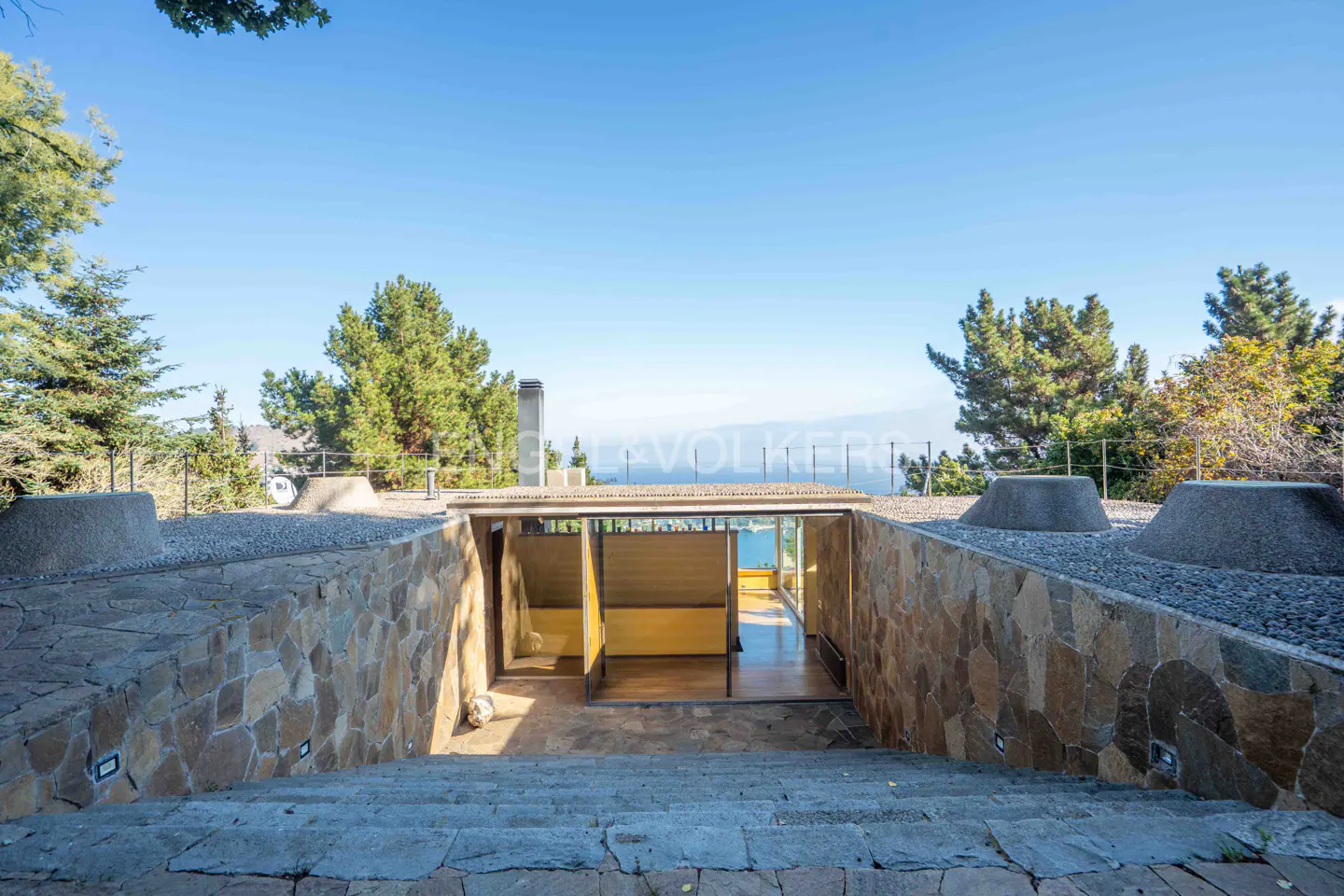 Stone steps lead down to a modern home entrance with glass doors, surrounded by stone walls and lush trees under blue sky.