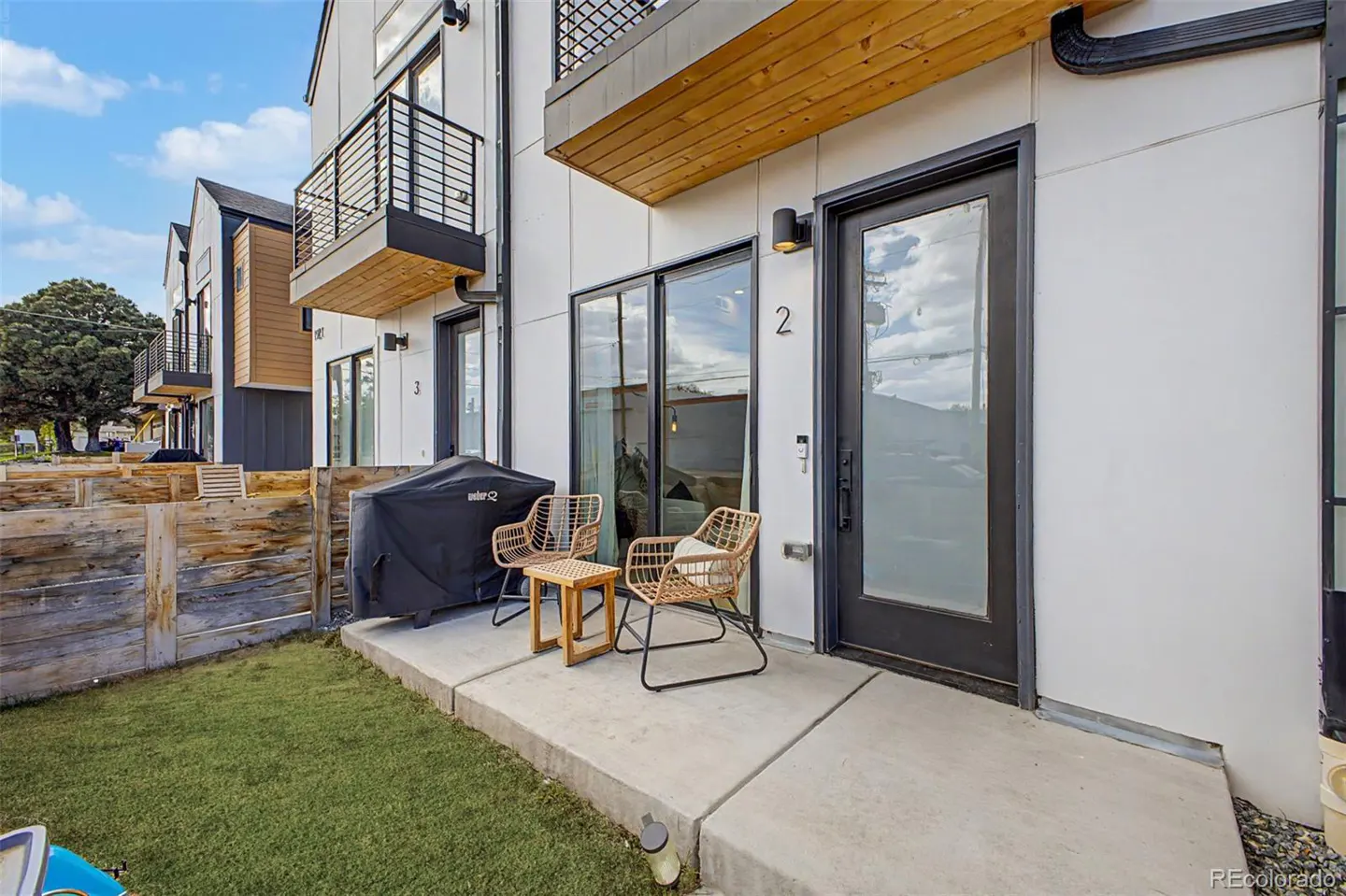 Exterior view of a modern townhouse with a patio, grill, and seating. The house is white with black trim and a wooden accent. The patio has two chairs and a small table.