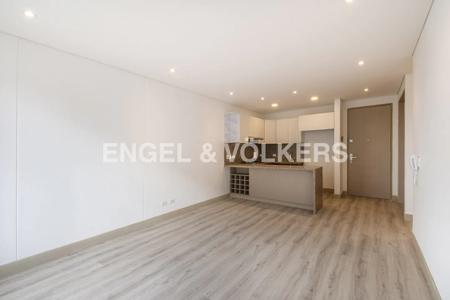 An open-concept apartment with light wood floors, white walls, and a kitchen island. The kitchen has white cabinets and a wine rack.