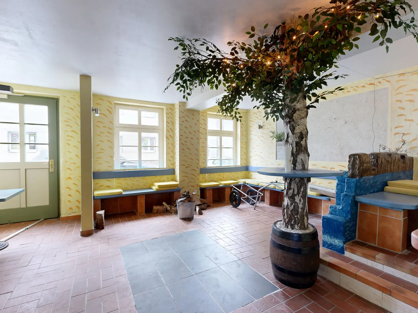 Interior view of a room with a tree growing from a barrel, benches, and a brick floor. The walls are yellow with a pattern.