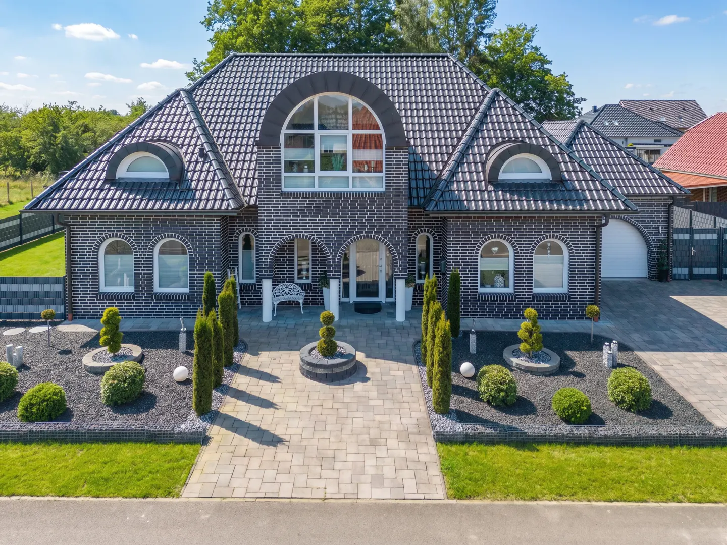 Two-story brick house with arched windows and a gray tiled roof. A stone walkway leads to the front door, surrounded by manicured landscaping.