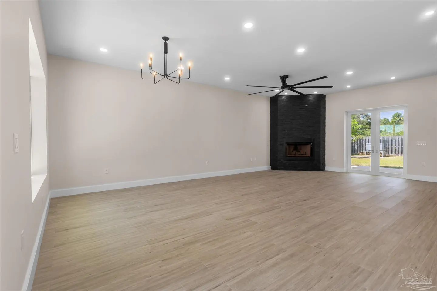 A spacious living room with light wood floors, a black brick fireplace, and a modern chandelier. Sliding glass doors lead to a green backyard.