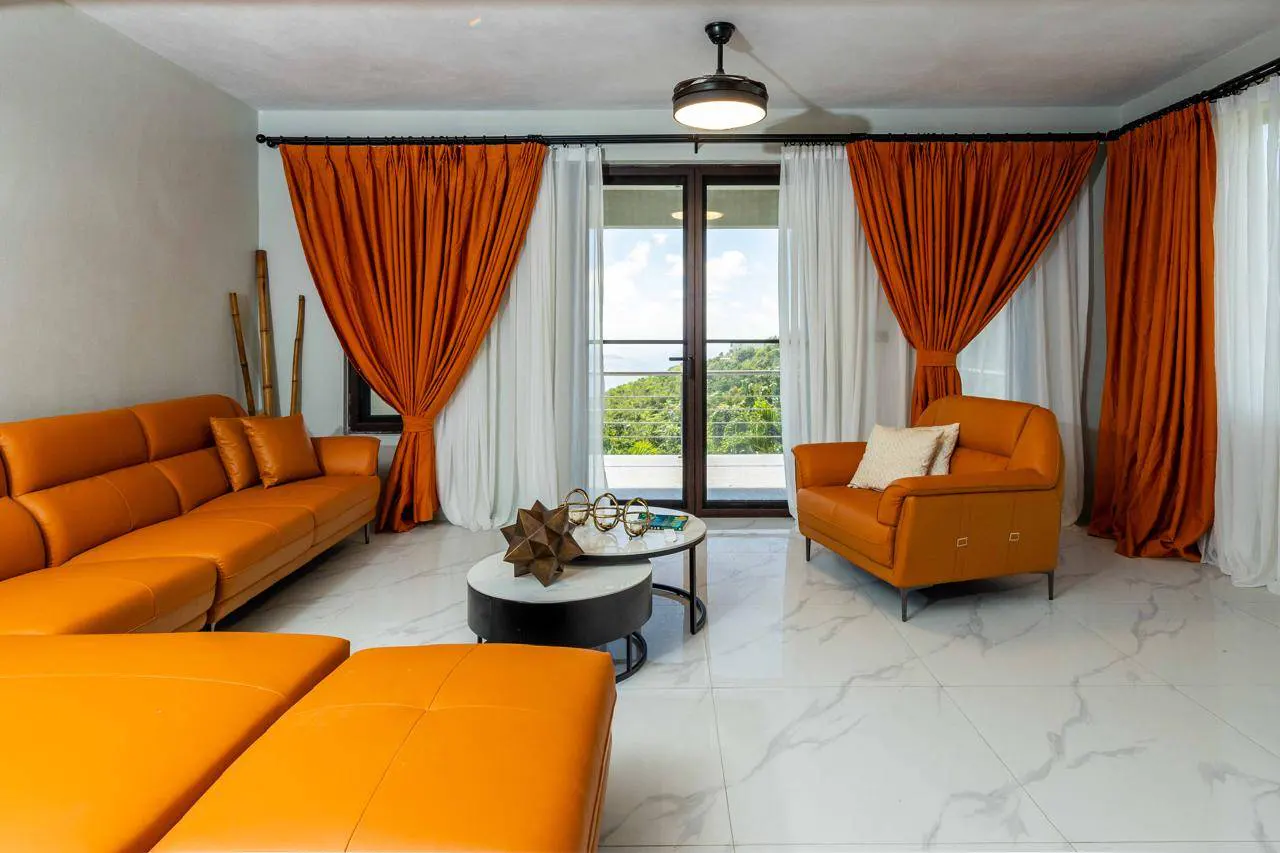 Bright living room with orange leather sofa and chair, white marble floors, and orange and white curtains. Balcony view of lush greenery.