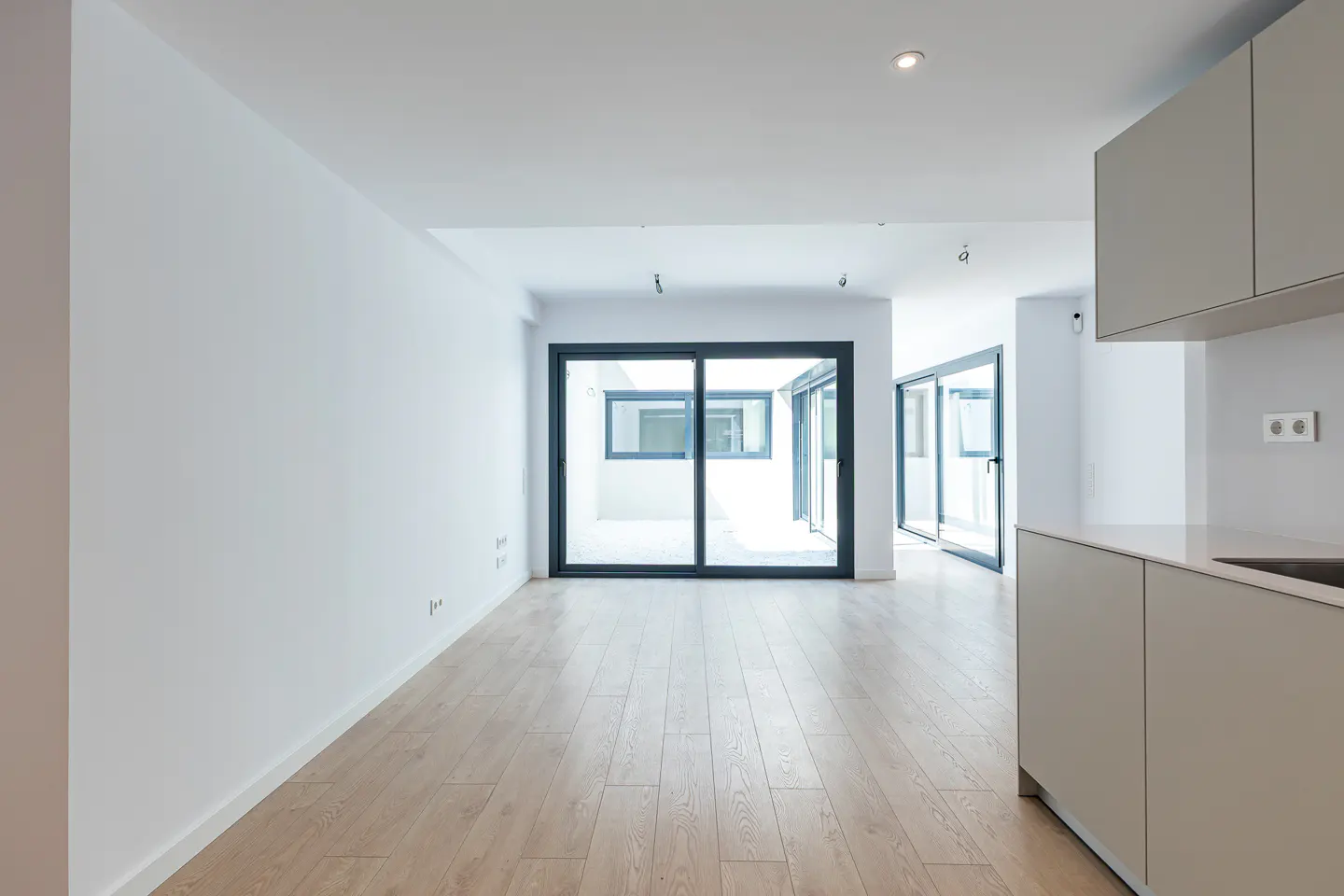 Bright, empty room with light wood floors, white walls, and sliding glass doors to a patio. Modern gray kitchen cabinets are on the right.
