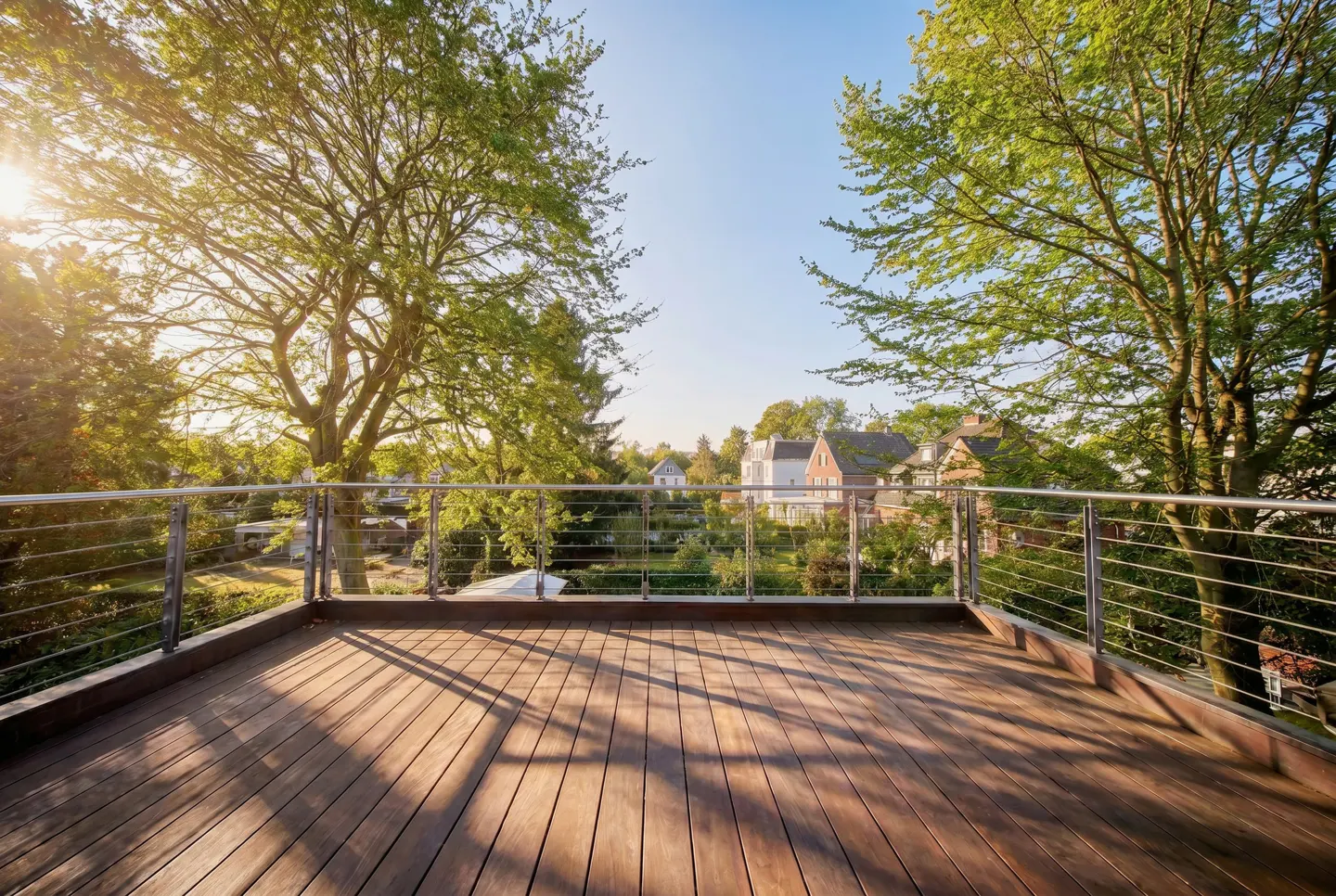 Wooden deck with metal railing overlooking a neighborhood with green trees and houses under a blue sky.