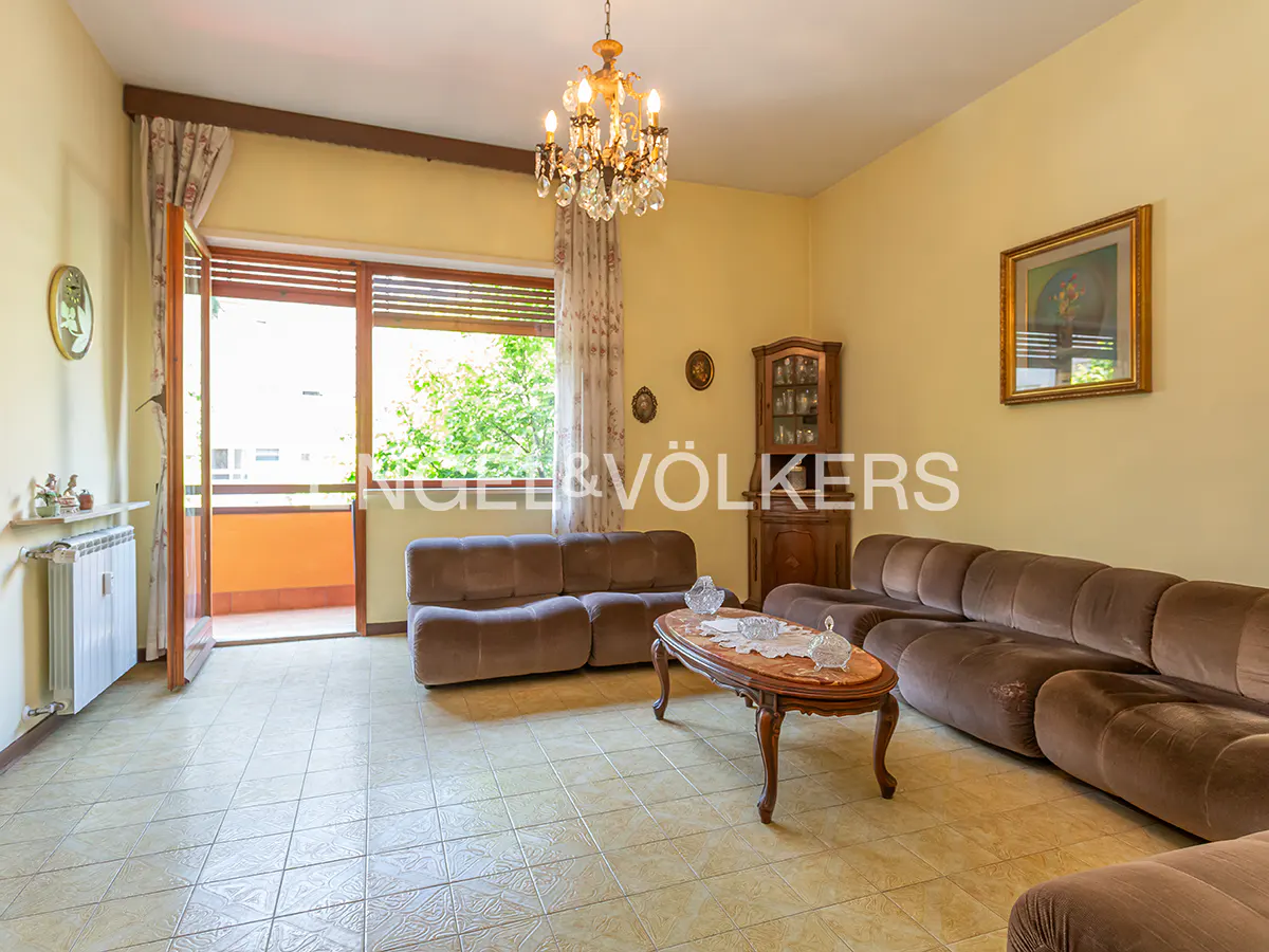 Living room with brown modular sofas, oval wooden coffee table, crystal chandelier, and balcony door with garden view.
