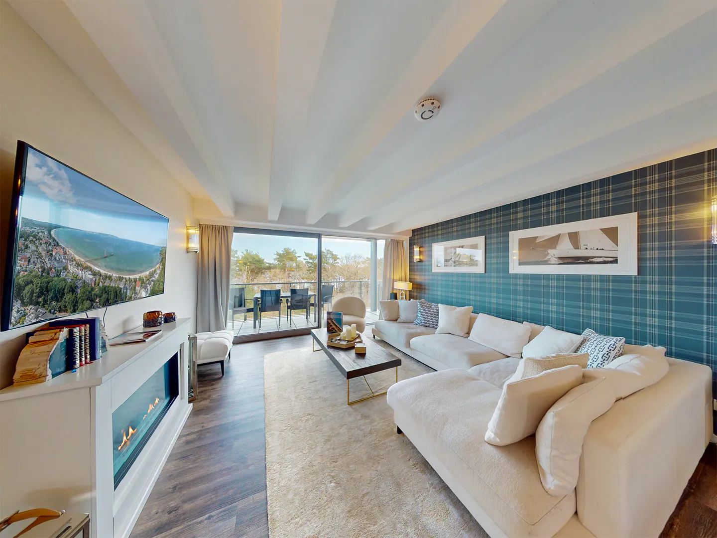 Living room with a large white sectional sofa, a TV showing a beach view, and a balcony with outdoor seating. The wall has blue plaid wallpaper.