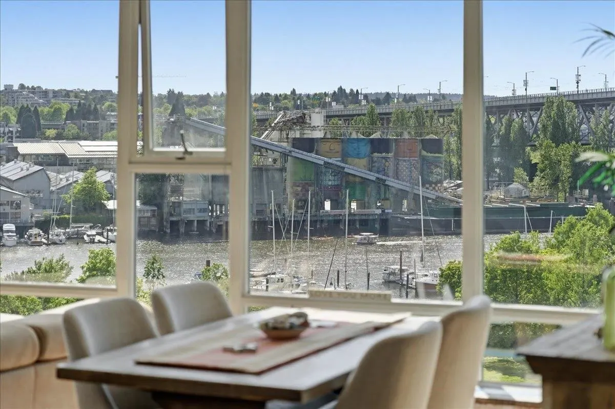 View from a dining room window overlooking a river with boats, a bridge, and industrial buildings. The table is set with a runner and bowl.