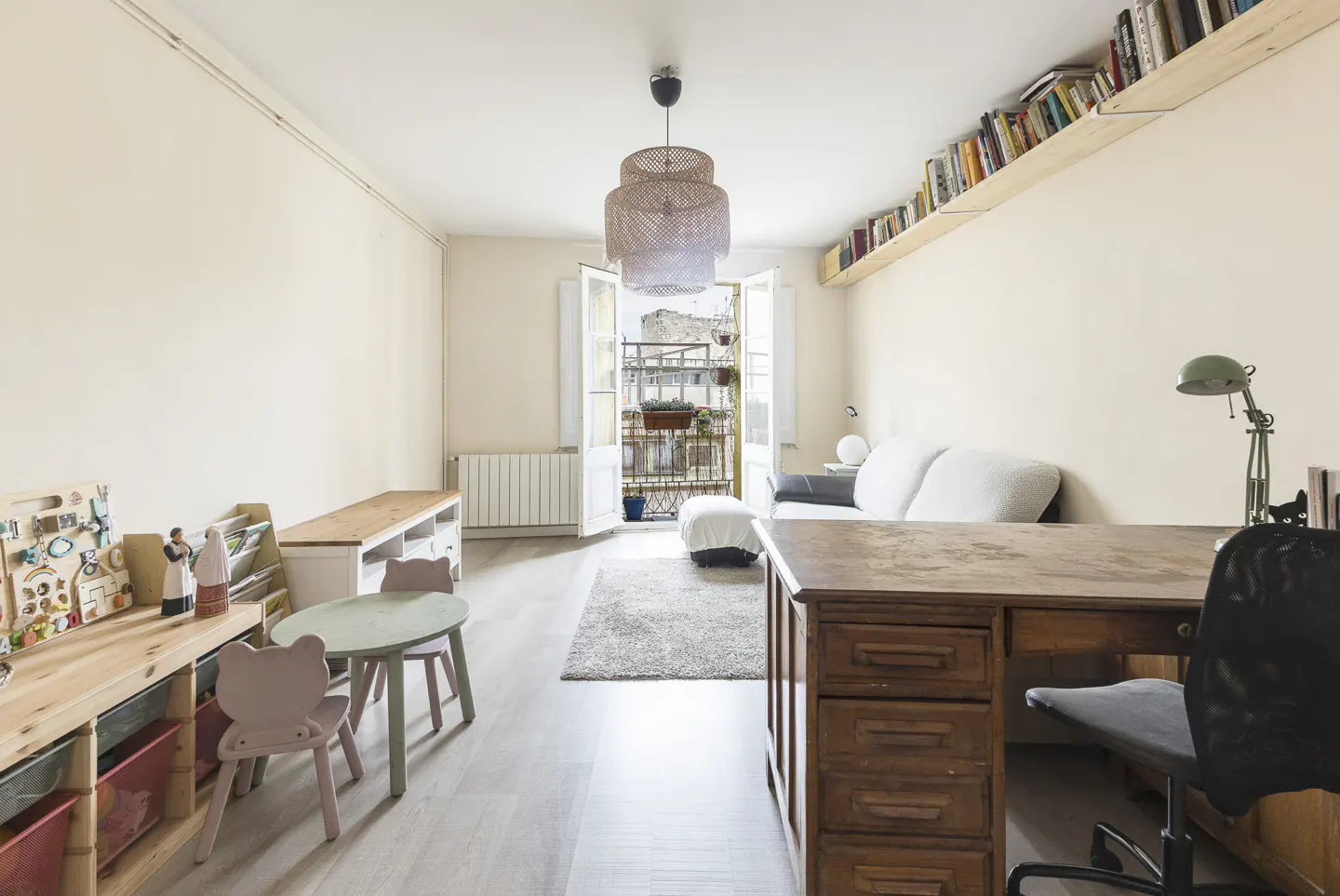 Bright living room with a desk, sofa, and children's play area. Balcony doors open to a city view. Bookshelf above the sofa.
