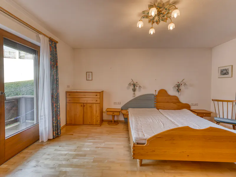 Bedroom with light wood furniture, including a bed, dresser, and nightstands. A door leads to a balcony with a view of a green hill.