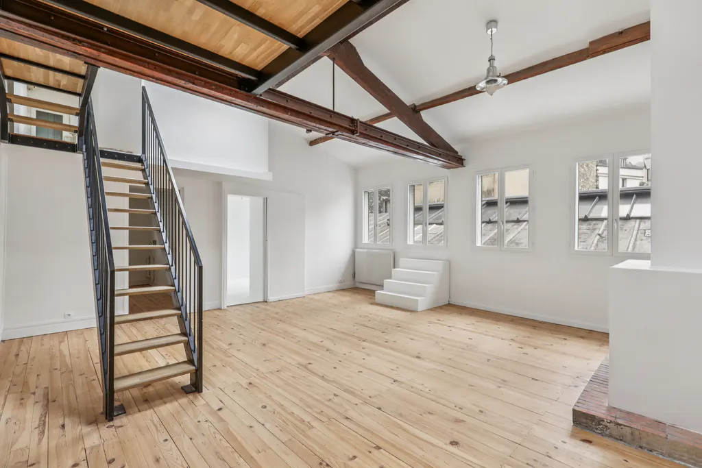 Bright, airy loft with wood floors, white walls, and exposed beams. A black metal staircase leads to a second level. Windows offer city views.