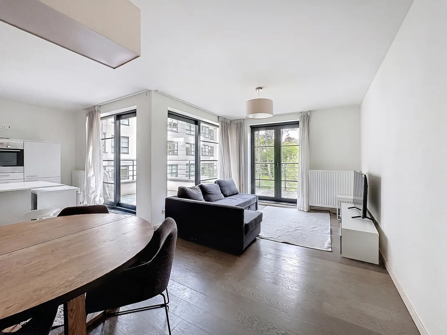 Bright, modern apartment with dark wood floors, a gray sofa, and a wooden dining table. Large windows offer outdoor views.