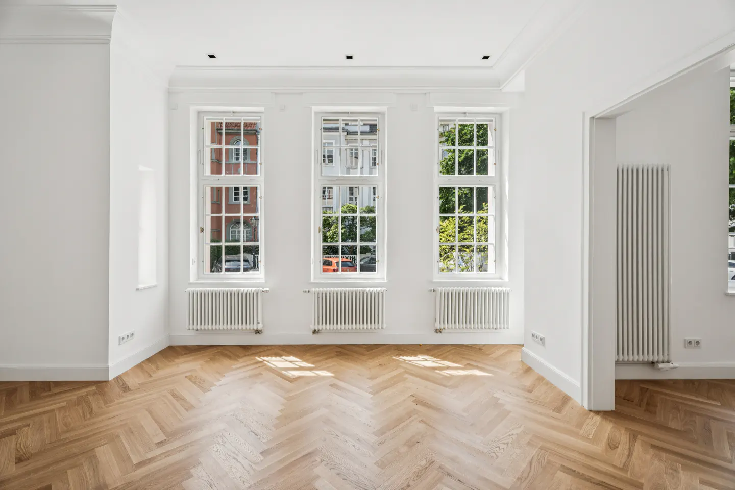 Bright, empty room with herringbone wood floors, white walls, three windows, and white radiators.