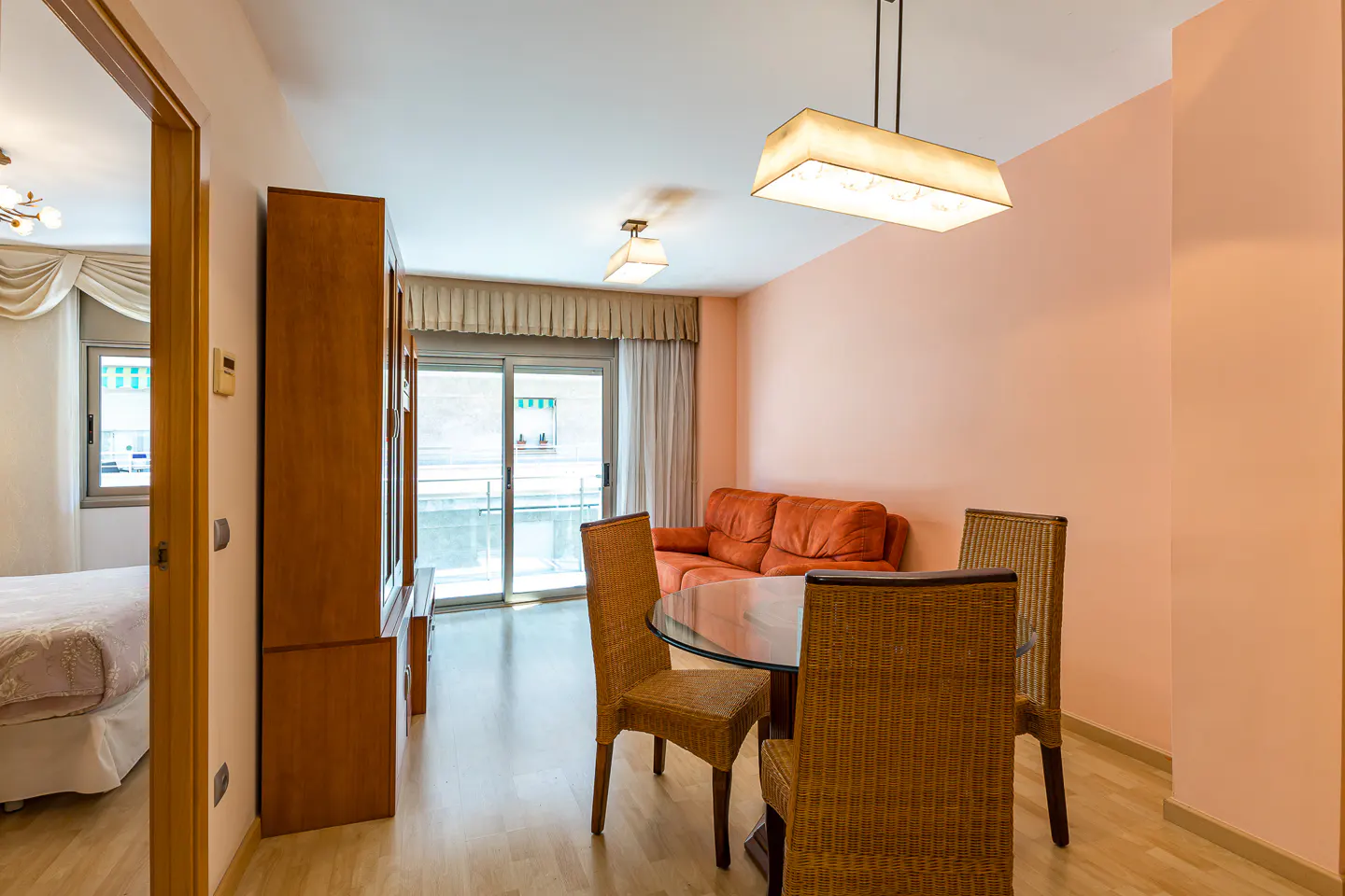 Dining area with a glass table and wicker chairs, orange sofa, and view into a bedroom with light wood floors.