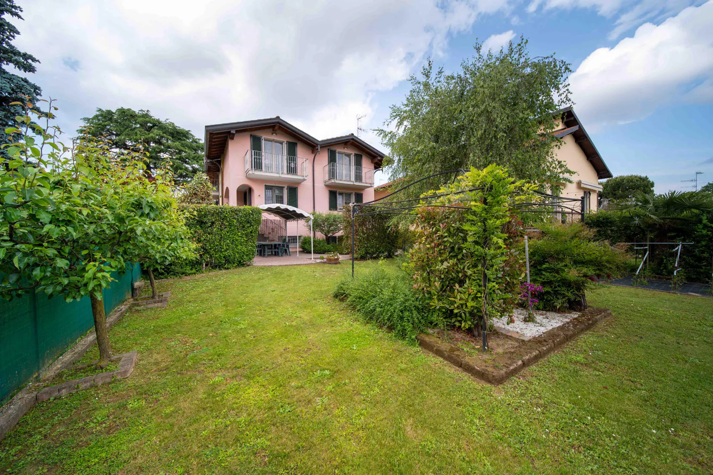 A two-story pink house with green shutters and balconies, surrounded by a green lawn and trees.