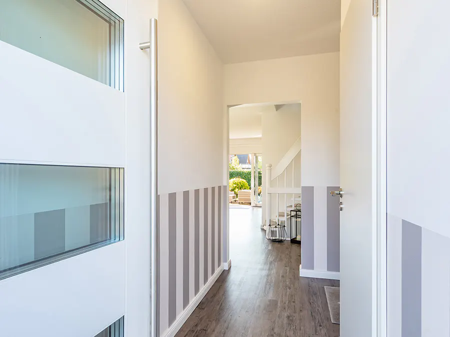 Open white door reveals a hallway with gray striped walls and wood-look floors. A staircase and outdoor view are visible.