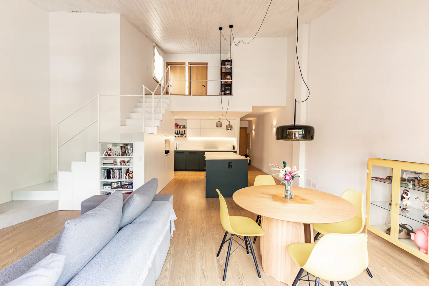 Modern open-plan apartment with a grey sofa, round wooden dining table, yellow chairs, and a loft staircase.