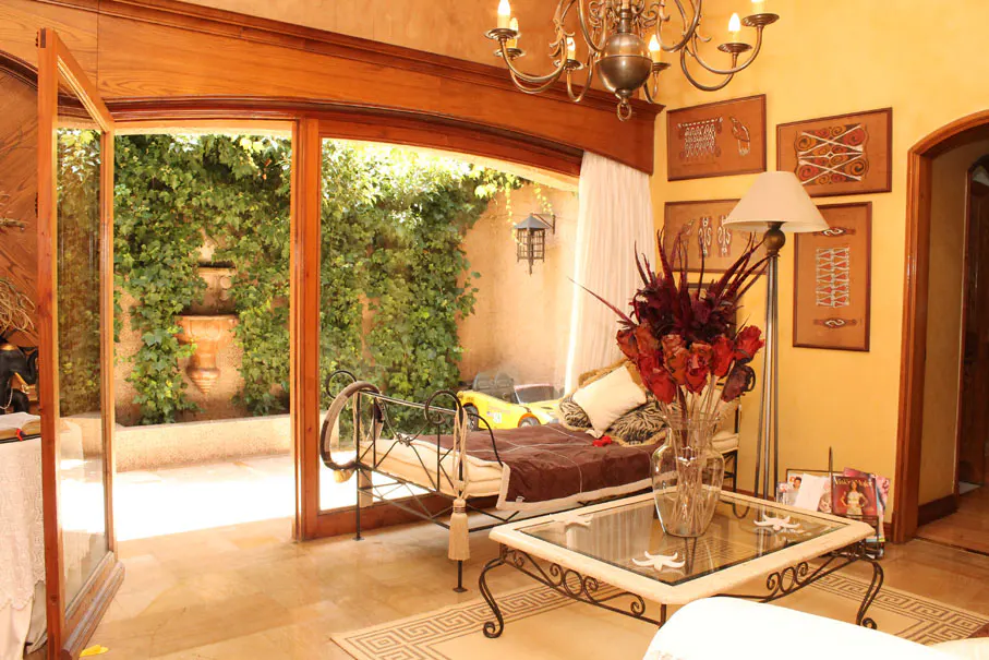 A sunlit living room with an open door to a courtyard. A daybed sits near the door, and a glass coffee table is in front of it. A chandelier hangs from the ceiling.